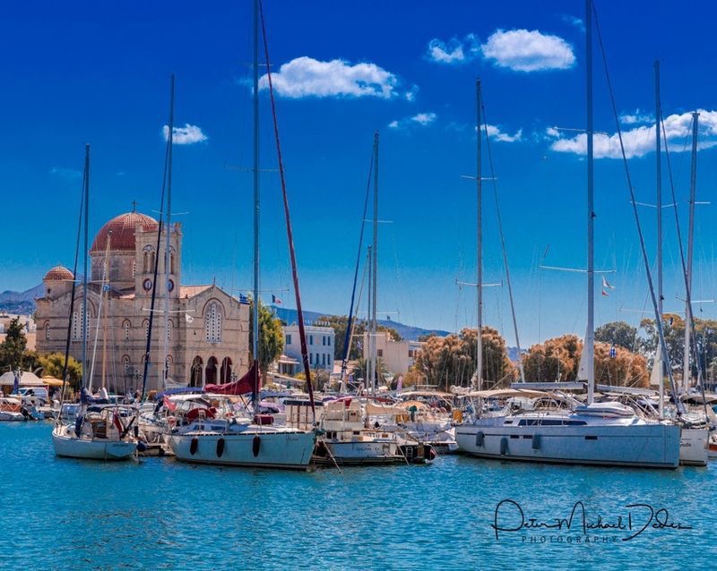 Sailboats docked in a Mediterranean harbor with a historic church and clear blue sky in the background.