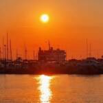 The sun sets over a busy harbor filled with boats and yachts, casting a warm golden glow over the water.