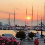 The sun sets over a bustling marina filled with sailboats, casting a warm glow on the water and the surrounding area.