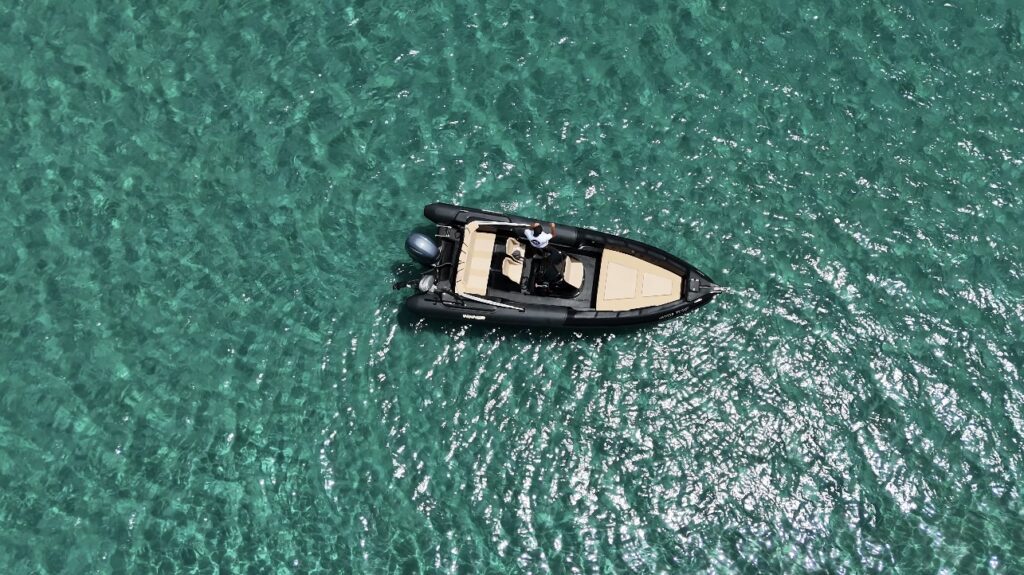 Overhead view of a compact black RIB boat with tan seating cruising on crystal-clear water.
